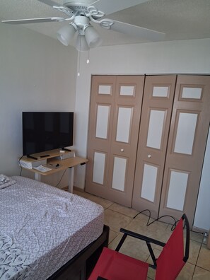 A bedroom with a bed, a desk with a TV, a ceiling fan, and a wardrobe.