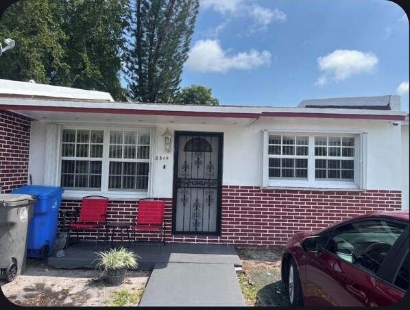 Une maison de plain-pied avec une façade en briques, une porte d’entrée agrémentée d’un portail en métal décoratif, un petit porche avec des chaises rouges et une voiture rouge garée dans l’allée.