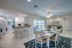 Una cocina moderna con armarios blancos, un comedor con mesa y sillas blancas, y un amplio ventanal con vistas a una piscina.