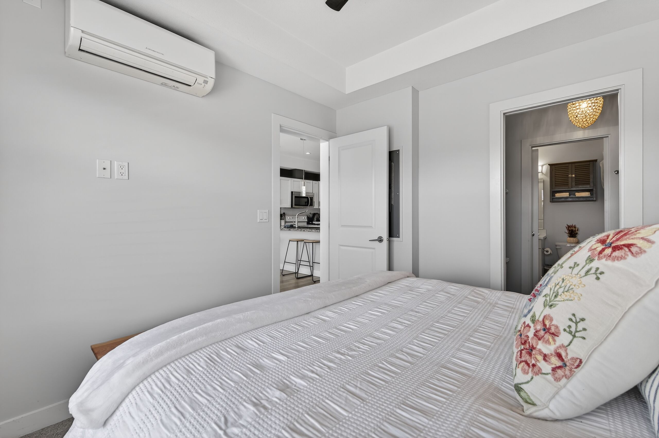 A bedroom with a bed, an air conditioning unit, a door leading to a kitchen, and a light fixture.