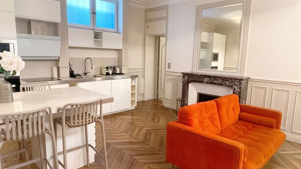 A modern kitchen with white cabinetry, a dining area with a white table and chairs, and a bright orange sofa. -  Le Flandrin Céline 24 (Paris)