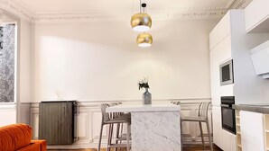A modern kitchen with a marble island, two pendant lights, and a dining area with chairs. -  Le Flandrin Céline 24 (Paris)