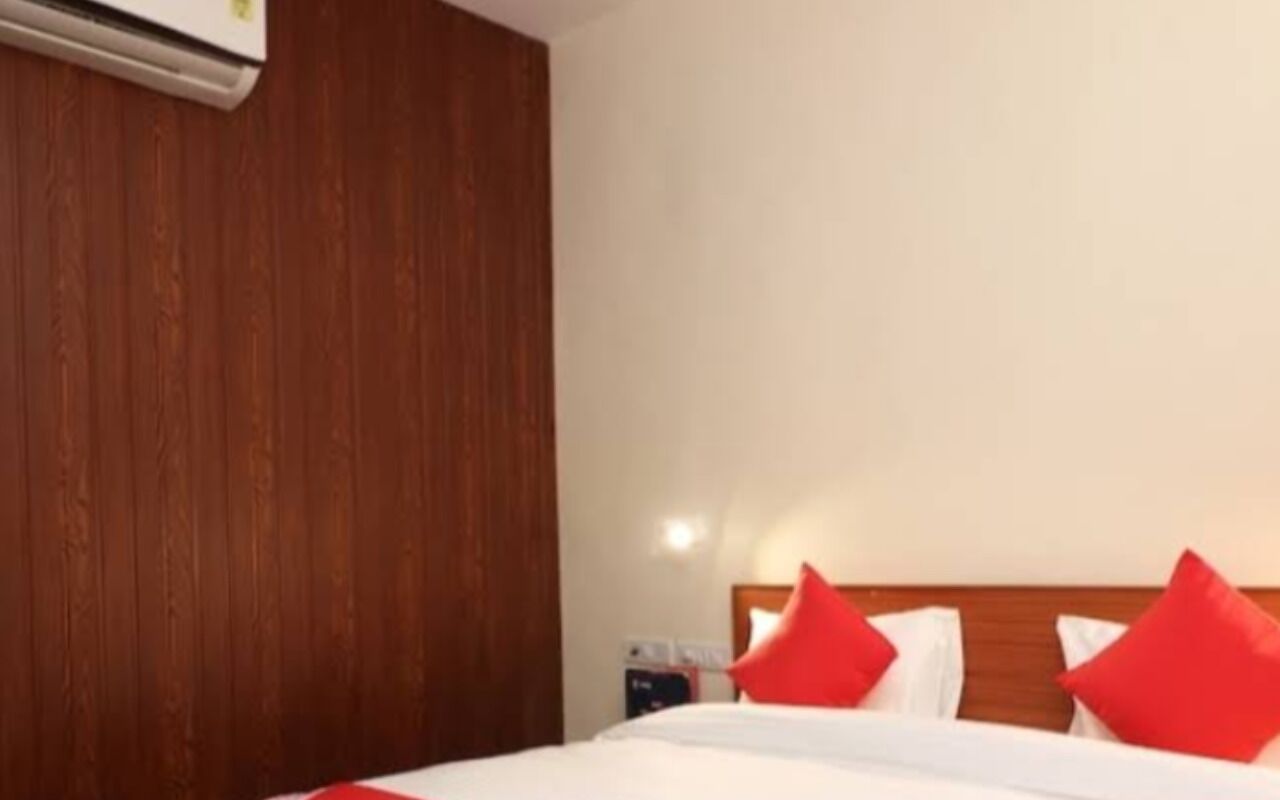 A hotel room with a wooden headboard, a bed with white bedding and red pillows, and an air conditioning unit mounted on the wall.