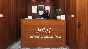 Un mostrador de recepción con la señalización 'HMI Hotel Meerut International', diplomas enmarcados en la pared y una planta en maceta.