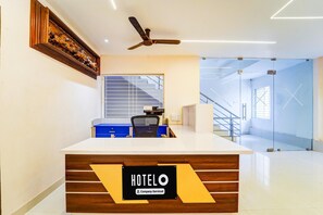 A hotel reception desk with a sign reading 'HOTELO Company-Serviced,' a ceiling fan, a blue office chair, and a staircase in the background. - Super Hotel O Aarapalayam (Madurai)