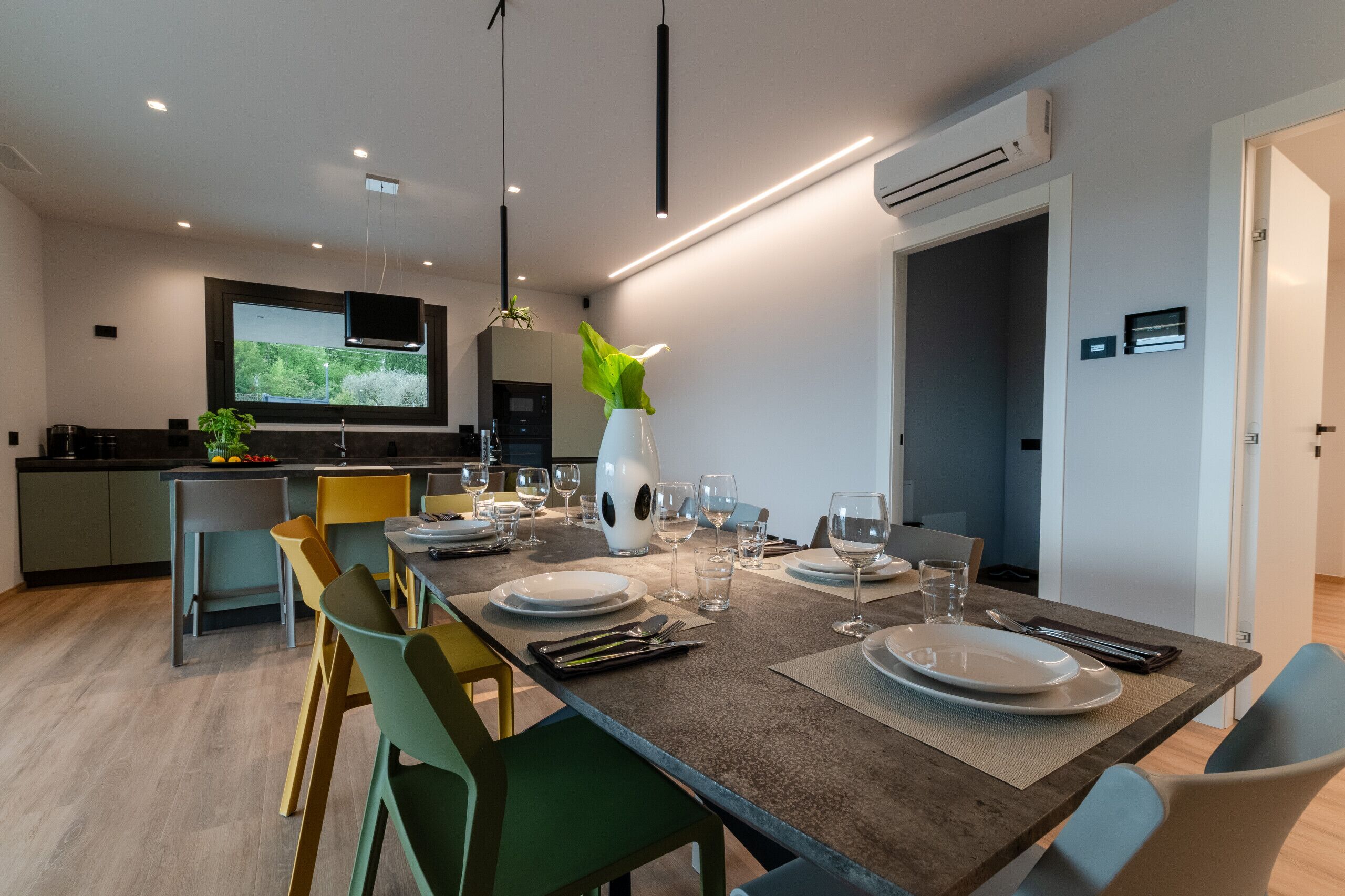 Uma sala de jantar moderna com uma mesa comprida posta para quatro pessoas, uma televisão fixada na parede e um aparelho de ar condicionado.