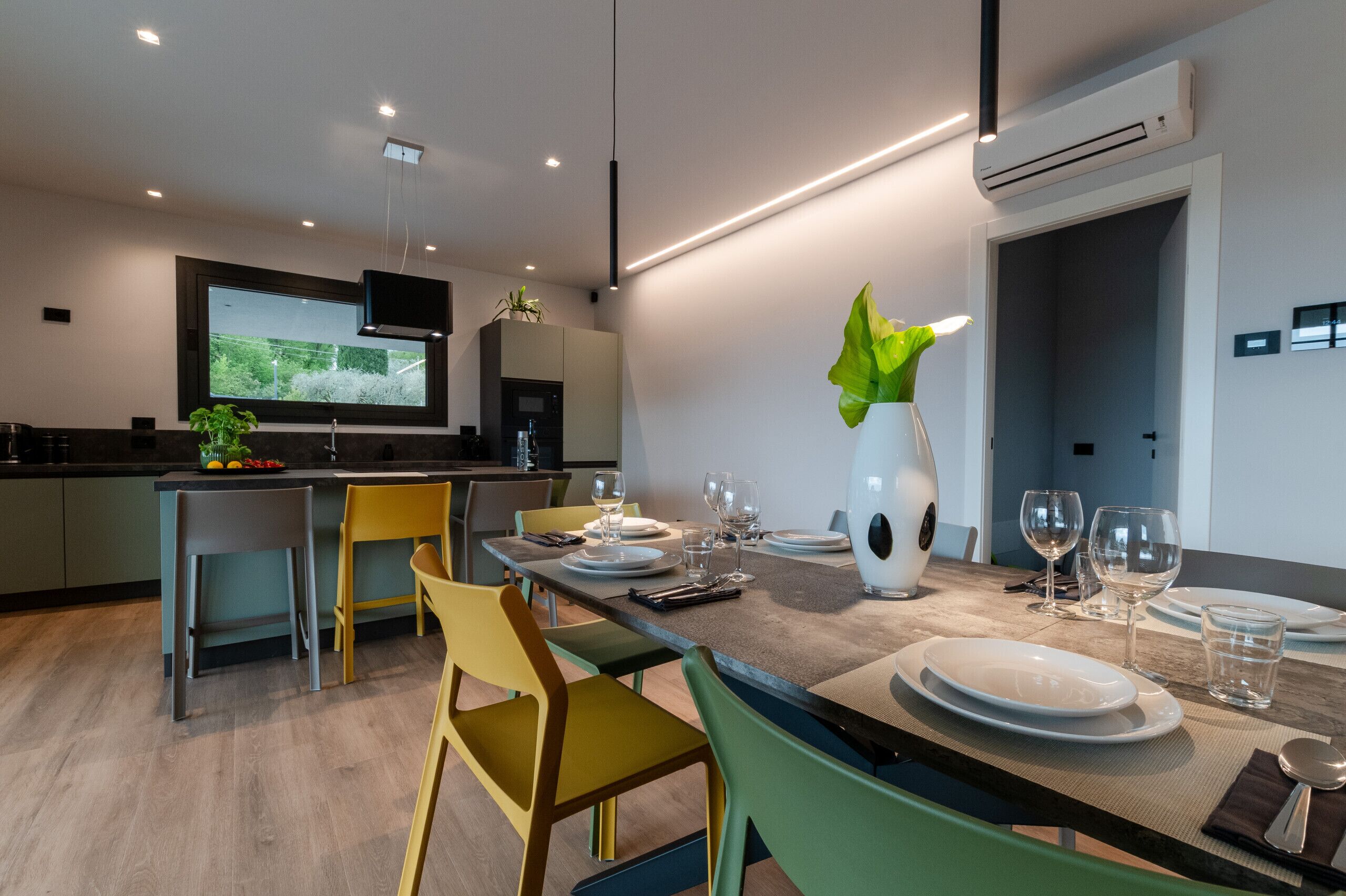 A modern kitchen with a dining area set for six, featuring a large flat-screen TV, a vase with a plant, and a dining table with chairs.