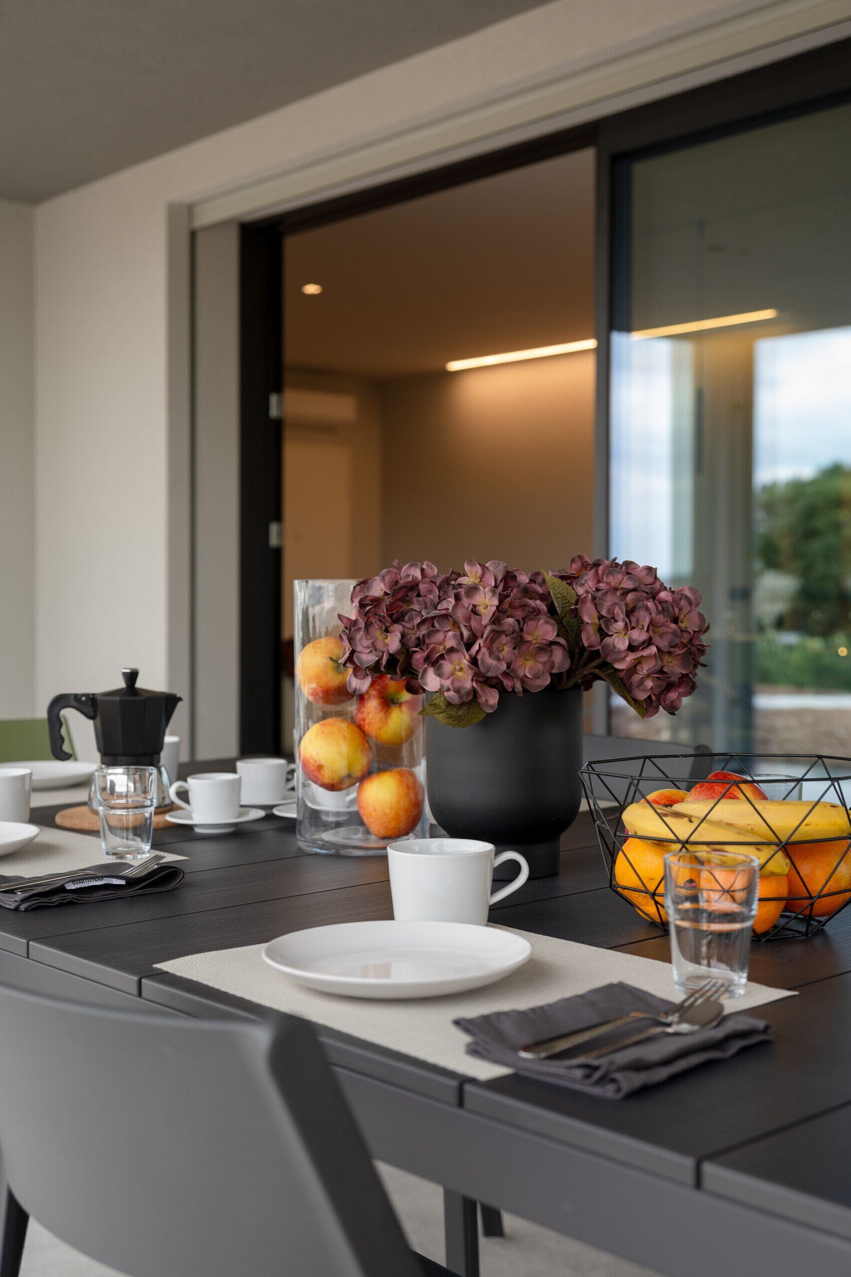 Una mesa de comedor dispuesta con platos, una cafetera, un florero con flores y una cesta de frutas.