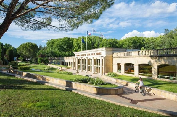 A building with columns and a flagpole, surrounded by a well-maintained garden and a paved walkway. - Les Hautes Plaines Residence with a pool open from 2:00 p.m. to 7:00 p.m. (Gréoux-les-Bains)