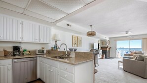 A modern kitchen with white cabinetry, a built-in dishwasher, and a large island with a sink. There is a dining area with a sofa and a view of the ocean. - Three Bedroom Two Bath Townhome Directly On The Bay (Ocean City)