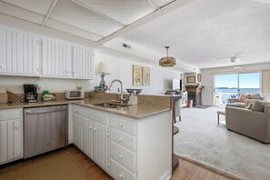 A modern kitchen with white cabinetry, a built-in dishwasher, and a large island with a sink. There is a dining area with a sofa and a view of the ocean. - Three Bedroom Two Bath Townhome Directly On The Bay (Ocean City)
