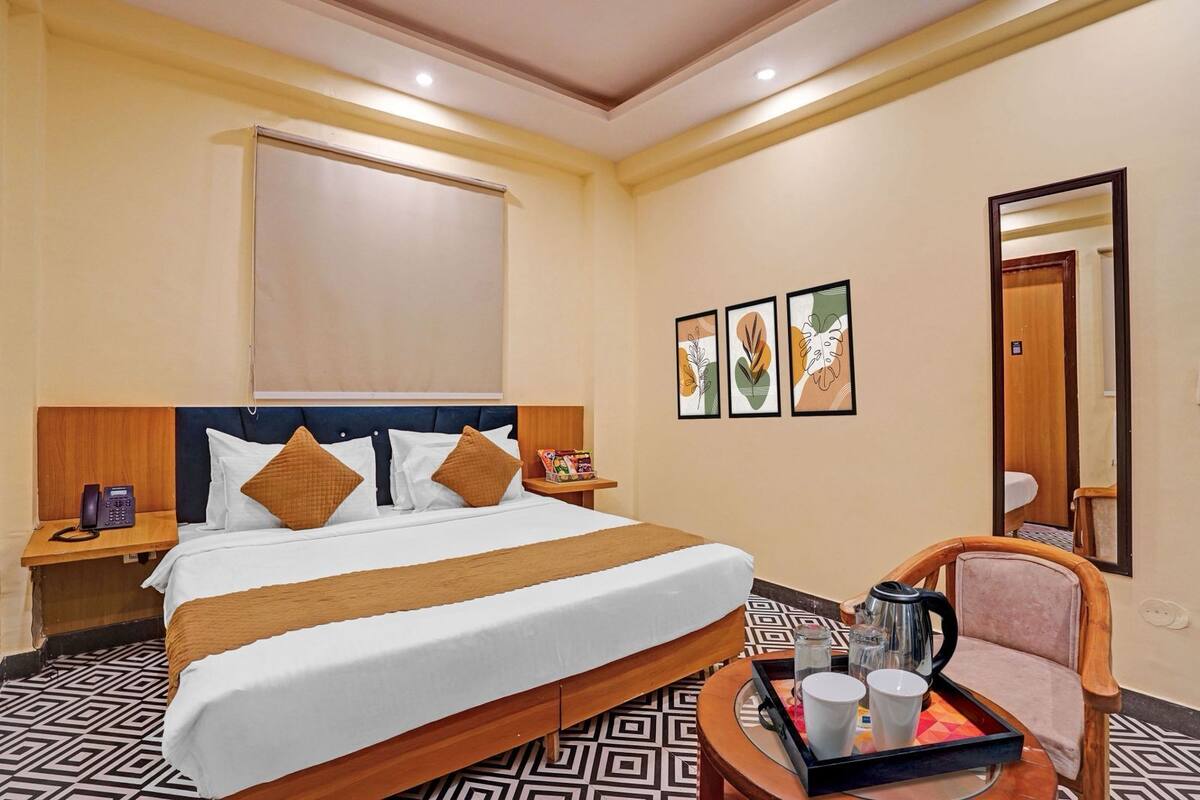 Quarto de hotel com uma cama grande, mesa de cabeceira com telefone, uma cadeira, bandeja com xícaras e chaleira, e espelho na parede.