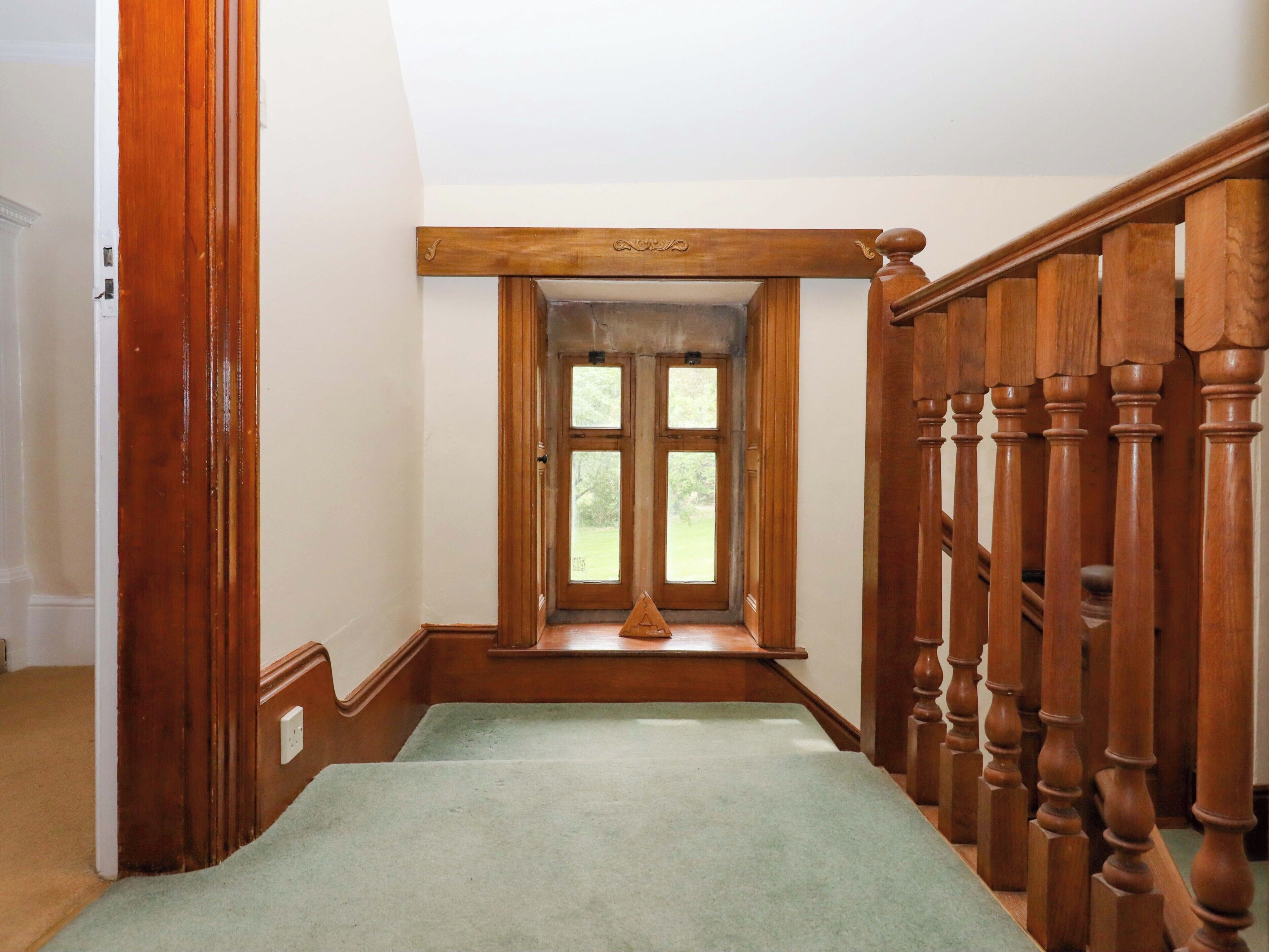 A wooden staircase with a green carpet, a wooden handrail, and a window with a view of greenery.