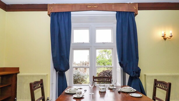 Une salle à manger avec une table en bois dressée pour quatre personnes, flanquée de deux chaises, une fenêtre avec des rideaux bleus et un radiateur au mur.