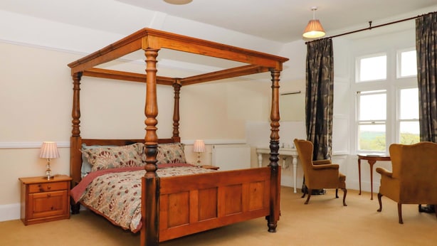A four-poster bed with a floral bedspread, two bedside tables with lamps, a chair, and a view of the outdoors through a window. - Dowrich West Wing Apartment (Crediton)