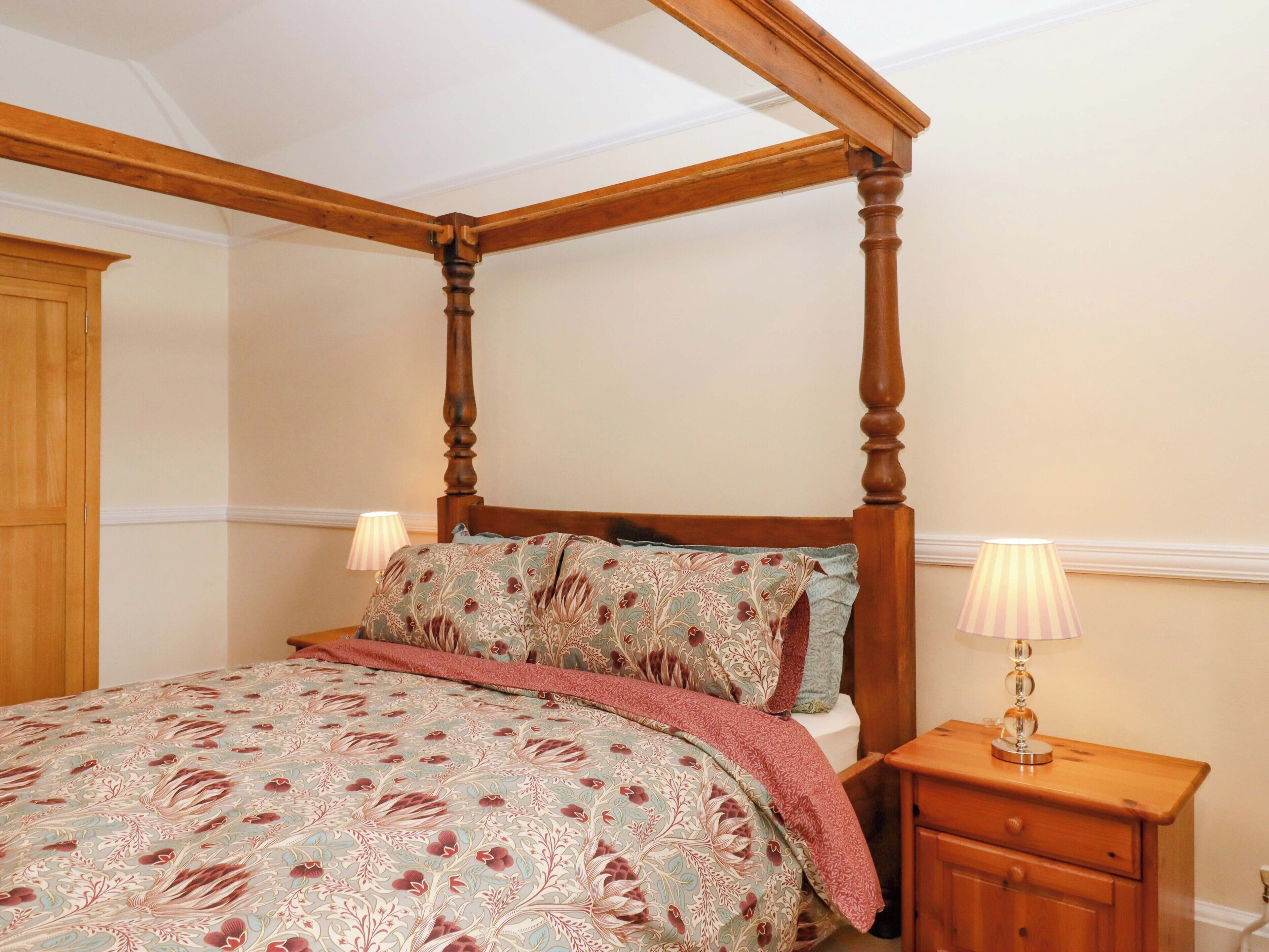 A four-poster bed with a floral bedspread, a nightstand with a lamp, and a wooden wardrobe.