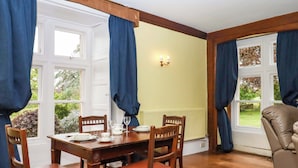 A dining room with a wooden table and chairs, a window with curtains, and a wall-mounted light fixture. - Dowrich West Wing Apartment (Crediton)