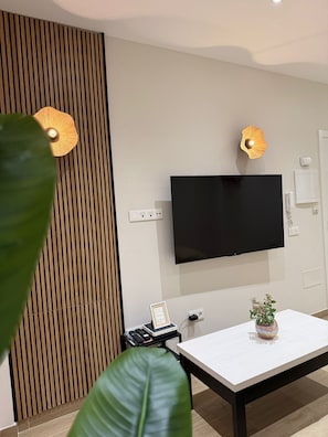 Ein modernes Wohnzimmer mit Flachbildfernseher an der Wand, einer Wandverkleidung aus Holzlamellen, einem Tisch mit Topfpflanze und einem kleinen Beistelltisch mit gerahmtem Gegenstand.