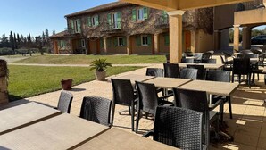 An outdoor patio area with tables and chairs, a building with green shutters in the background, and a grassy lawn. - Traveling in the 21st century is all about comfort (Mouriès)