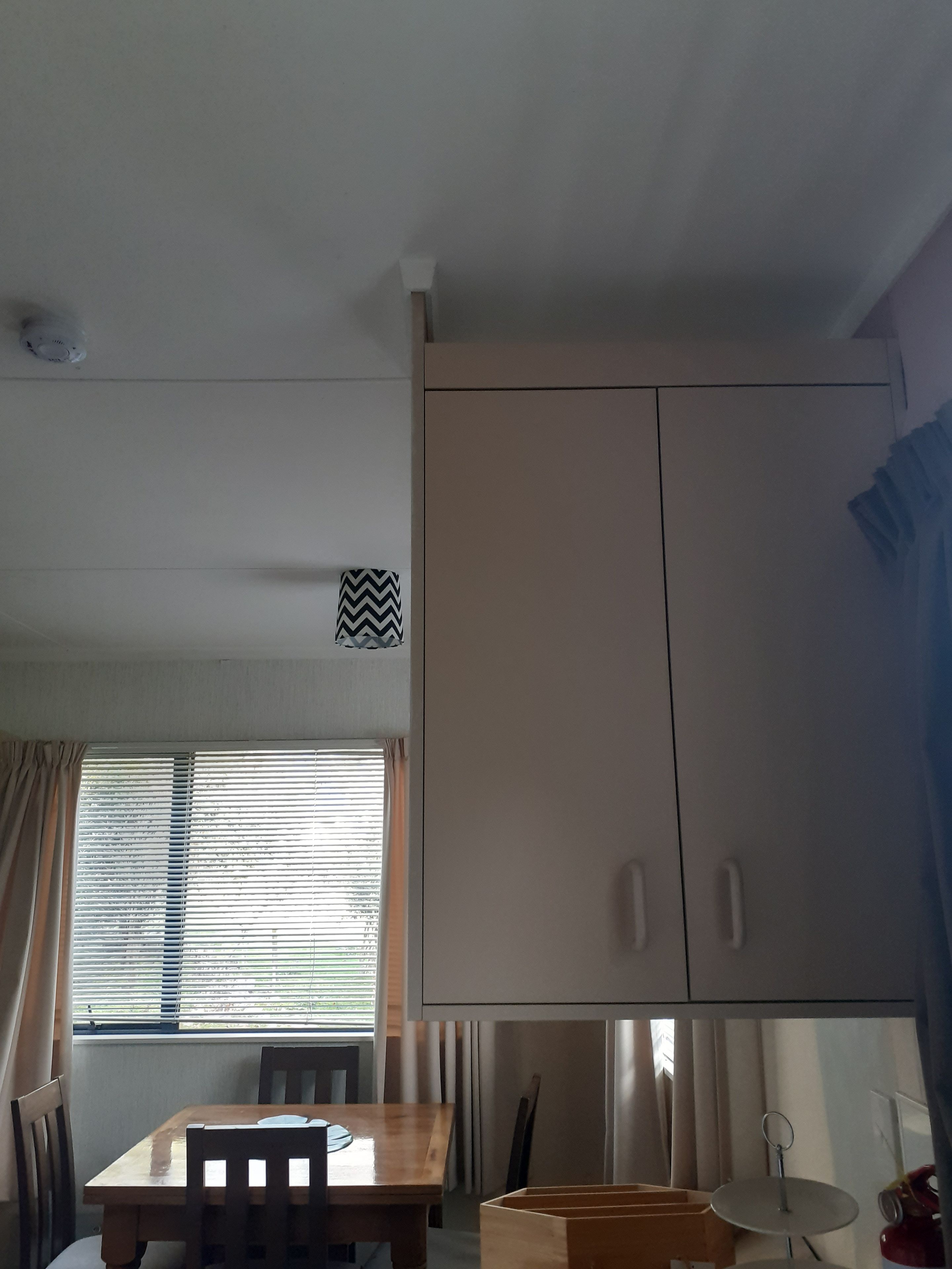 A room with a wooden table and chairs, a window with blinds, a ceiling light fixture, and a large wardrobe.