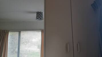 A room with a wooden table and chairs, a window with blinds, a ceiling light fixture, and a large wardrobe.