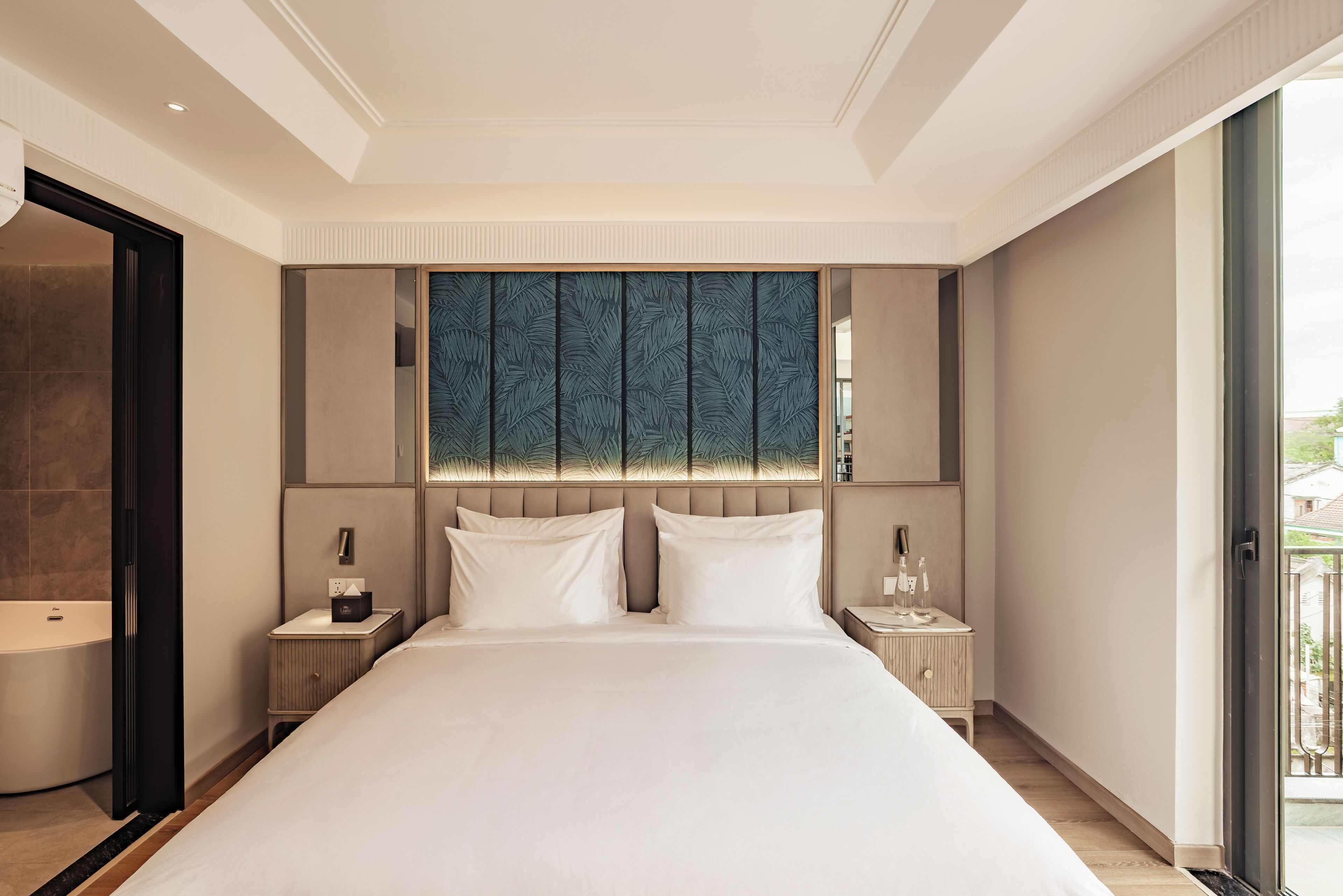 Une chambre d’hôtel moderne dotée d’un grand lit, d’une tête de lit intégrée, d’une table de chevet avec une lampe et d’une porte-fenêtre coulissante donnant sur le balcon.
