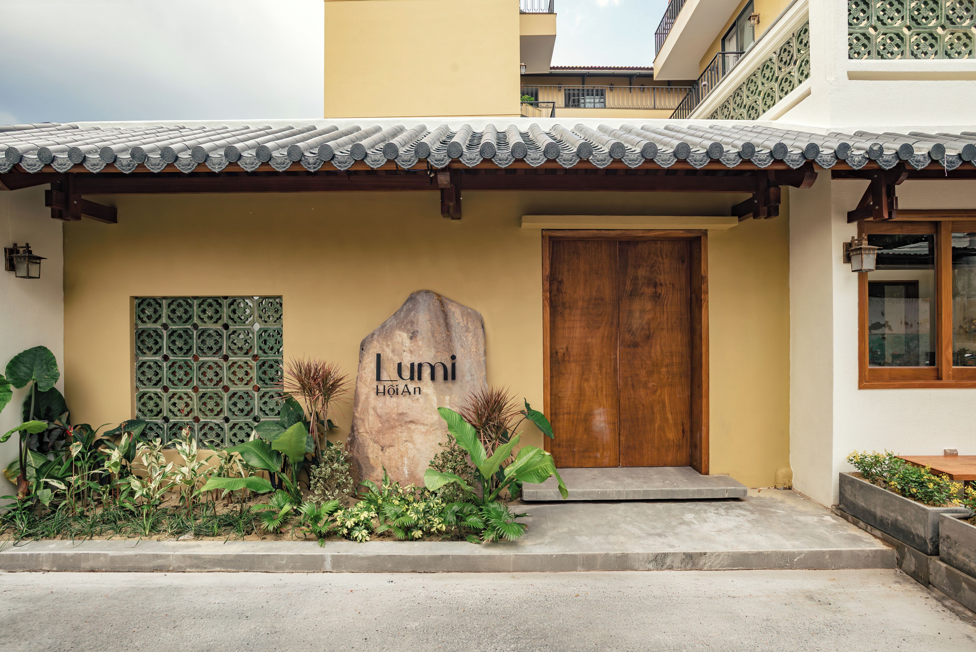 A building entrance with a tiled roof, a wooden door, and a large rock with 'Lumi HoiAn' carved on it.