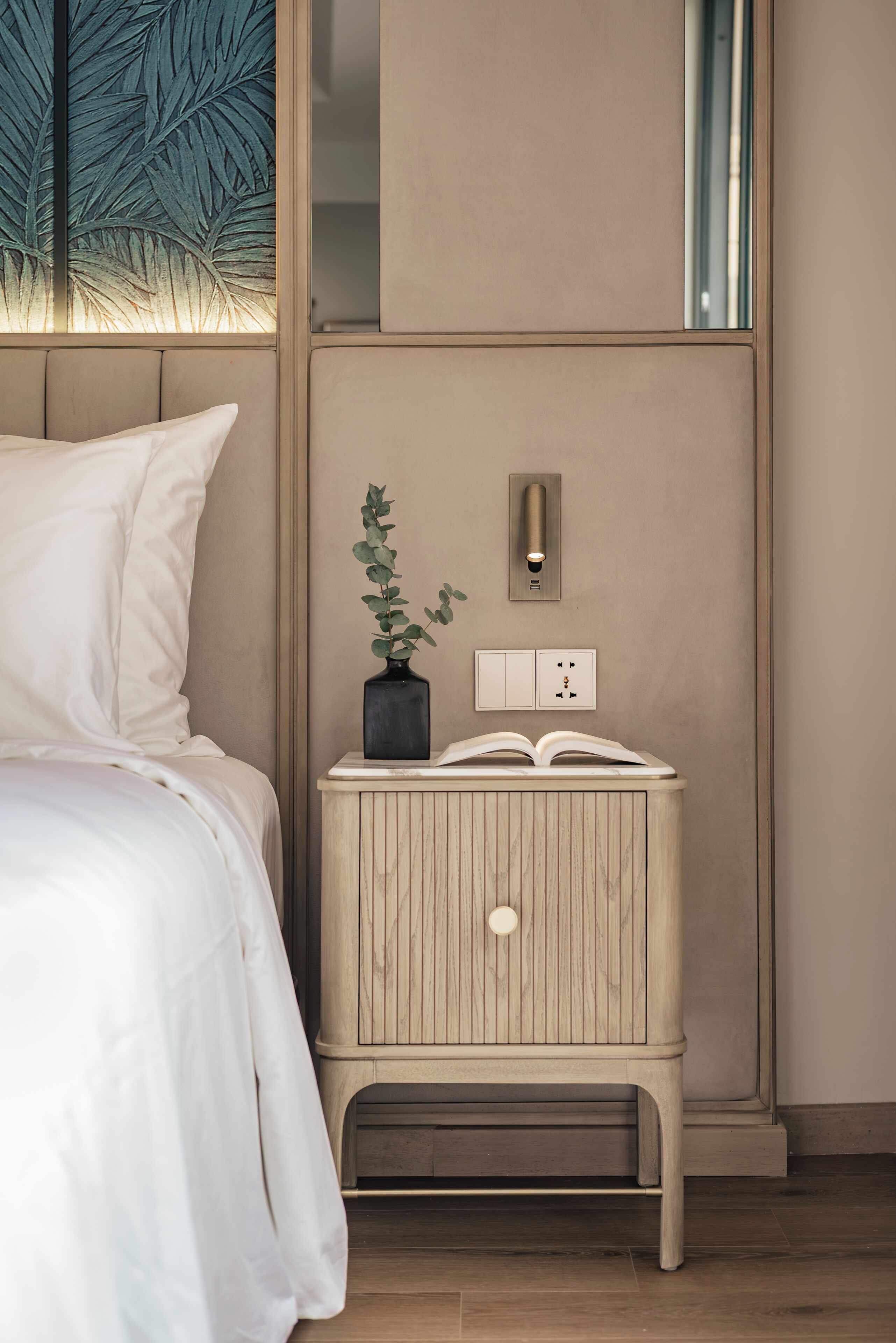 A bedroom with a bed, bedside table, a potted plant, a wall-mounted light, and a power outlet.