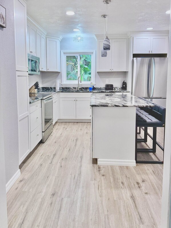 A modern kitchen with white cabinetry, a marble countertop, a stainless steel refrigerator, and a window with a view of greenery.