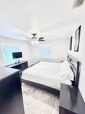 A bedroom with a bed, a desk, a ceiling fan, and two windows with blinds.