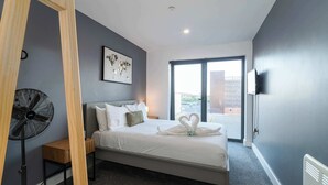 Et moderne hotelværelse med en seng, en standventilator, et lille trænatbord, et indrammet billede på væggen og et stort vindue med udsigt.