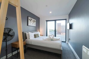 Una habitación de hotel moderna con una cama, un ventilador de pie, una mesita de noche de madera, un cuadro en la pared y un ventanal con vistas.