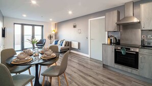 Et moderne køkken med indbyggede apparater, et spiseområde med et bord dækket til fire personer, og en opholdsstue med sofa og sofabord.