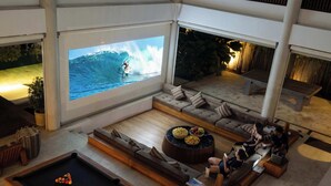 A modern living room with a pool table, comfortable seating, a coffee table with snacks, and a large TV screen displaying a surfer. - Luxury 9BR Bingin Villa - Large Pool, Near Beach (Kecamatan Kuta Selatan)