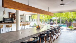 A modern kitchen with a long bar area, outdoor seating, and a view of a swimming pool. - Luxury 9BR Bingin Villa - Large Pool, Near Beach (Kecamatan Kuta Selatan)