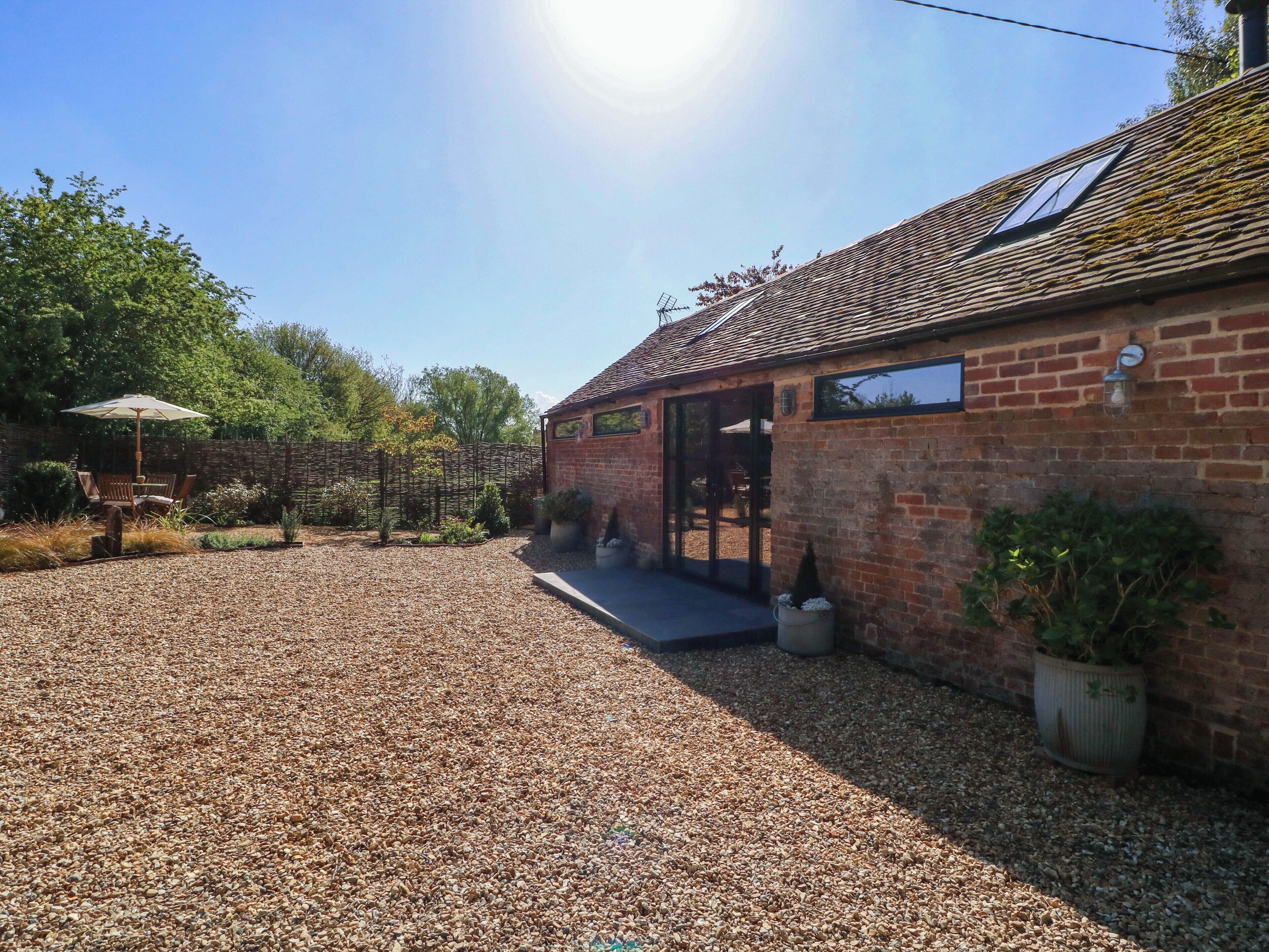 A brick building with a gravel driveway, a covered patio, and a garden area with plants and a wooden bench.