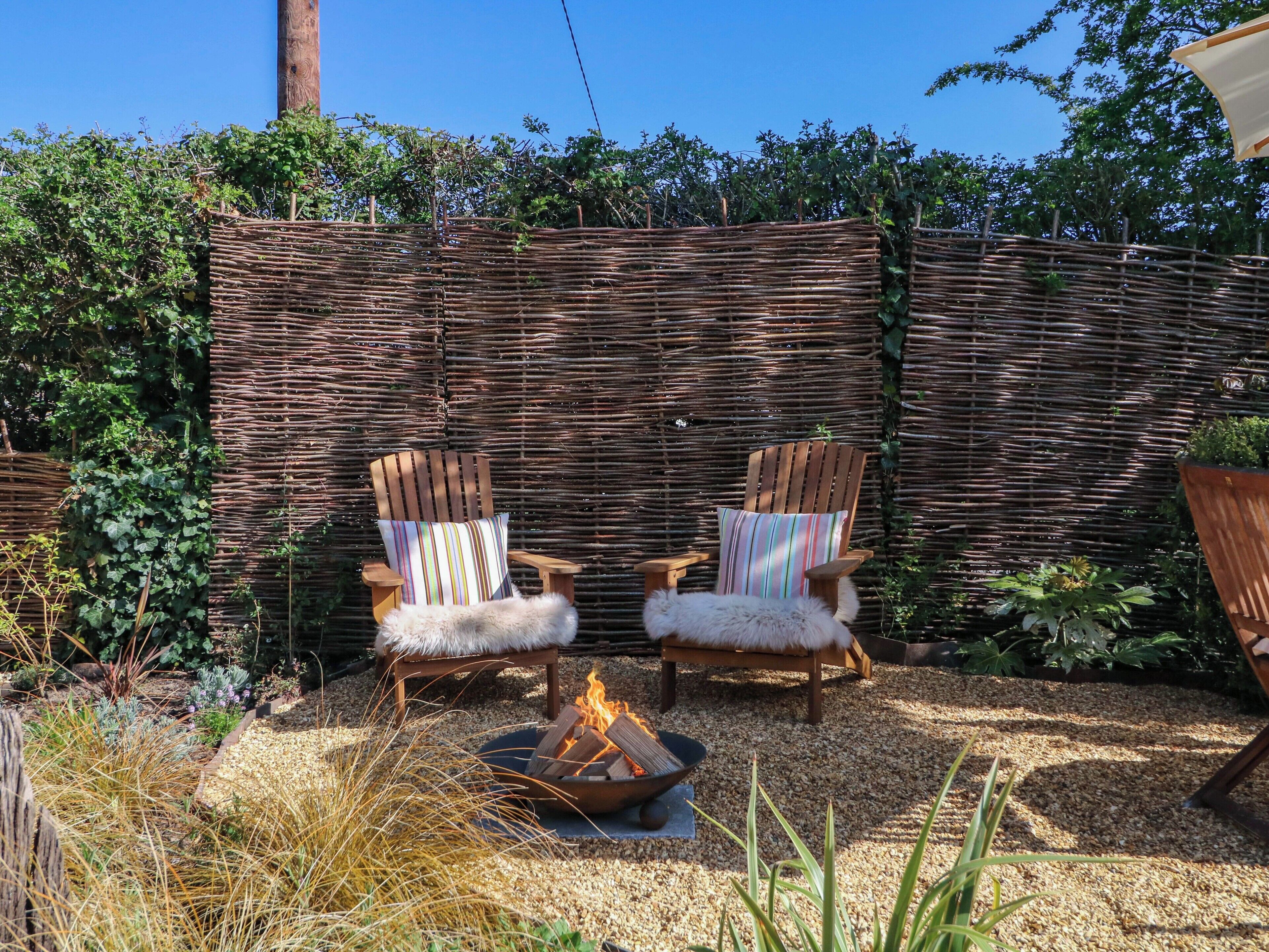 Two wooden chairs with striped cushions and fur throws, a fire pit, and a woven fence in a garden setting.
