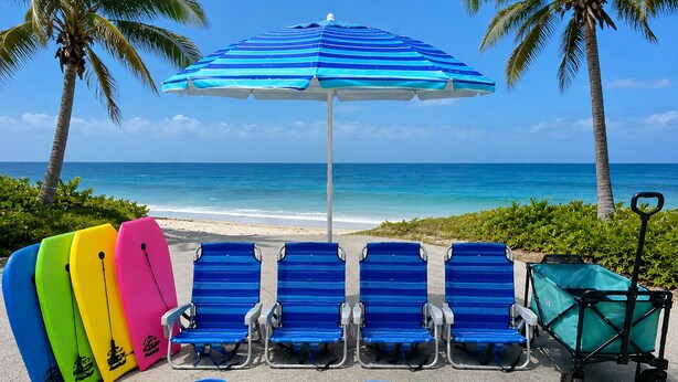 Una zona relax in spiaggia con poltroncine azzurre, un ombrellone a righe, giocattoli colorati e un carretto da spiaggia.