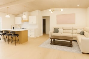 Eine moderne Küche mit weißen Schränken, einer zentralen Kochinsel mit Barhocker, einem Essbereich mit Sofa und Couchtisch sowie einem großen Gemälde an der Wand.
