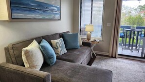 A living room with a gray sofa, a painting of a seascape, a window with blinds, a lamp on a side table, and a view of a balcony with outdoor furniture. - Cozy on the coast of Tybee (Tybee Island)