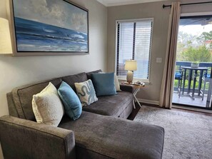 Un salon avec un canapé gris, un tableau représentant un paysage marin, une fenêtre avec des stores, une lampe sur une table d’appoint et une vue sur un balcon aménagé avec du mobilier d’extérieur.