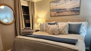 A bedroom with a bed, pillows, a bedside table with a lamp, a mirror, and a painting on the wall. - Cozy on the coast of Tybee (Tybee Island)