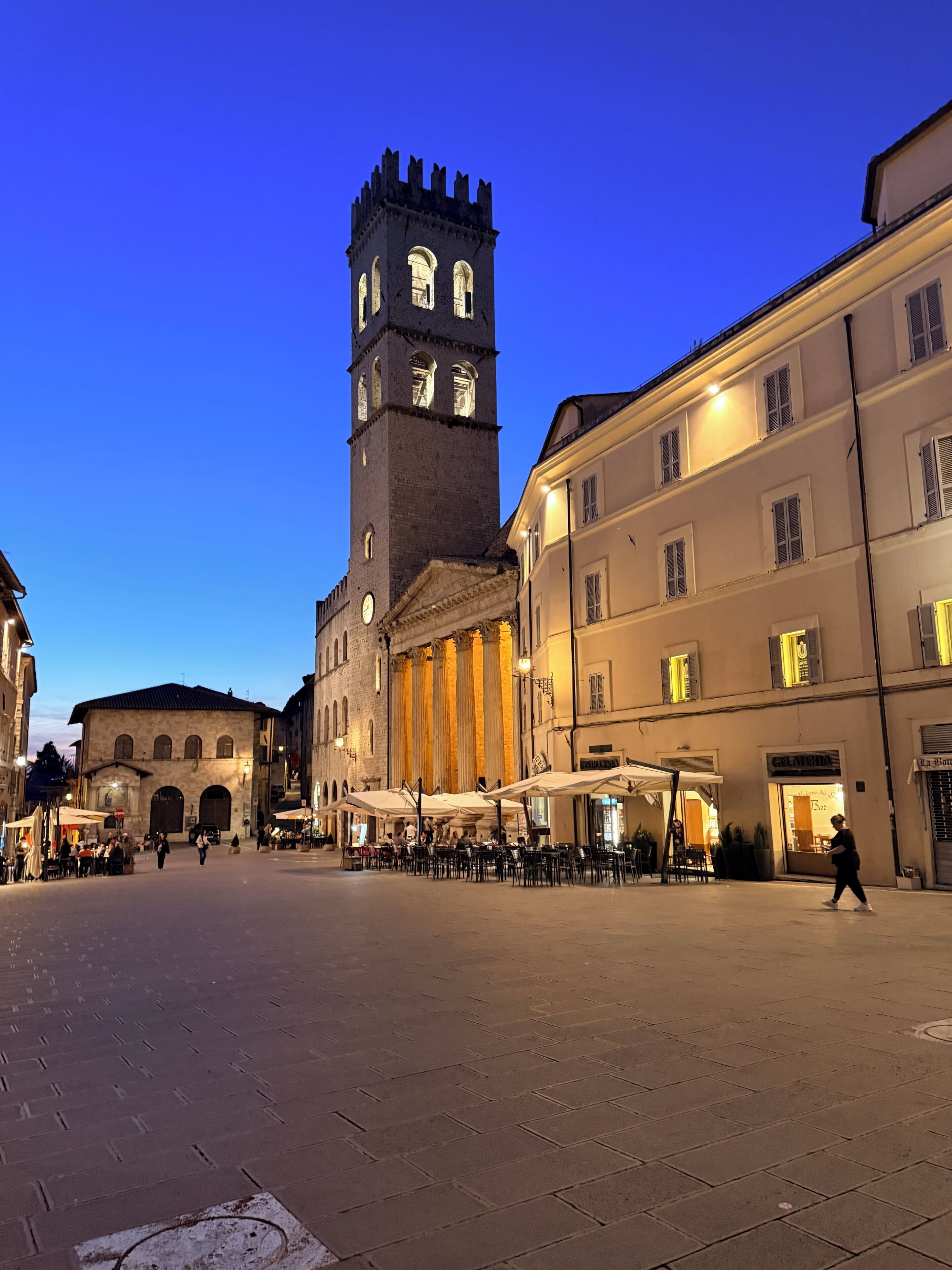 Una piazza storica con una torre imponente, posti a sedere all'aperto e un caffè.