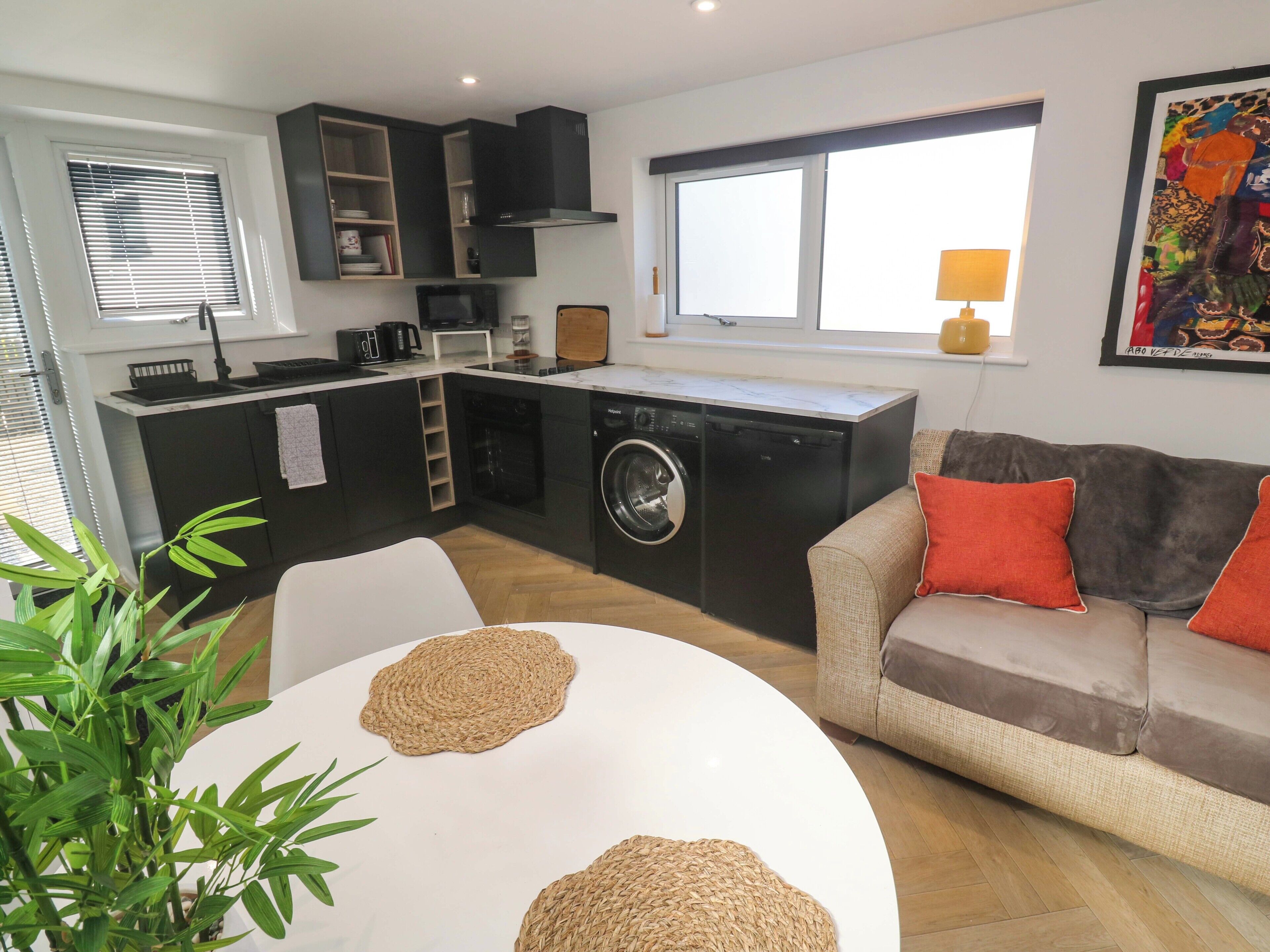 A compact living space with a kitchenette, a washing machine, a dining table with woven placemats, a sofa with cushions, and a large window.