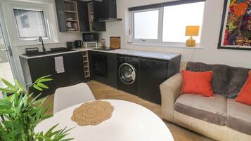 A compact living space with a kitchenette, a washing machine, a dining table with woven placemats, a sofa with cushions, and a large window.