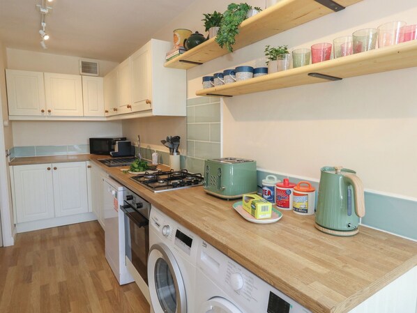 A kitchen with white cabinets, a washing machine, a microwave, a toaster, and shelves with kitchenware. - Mallow Cottage (Swanage)