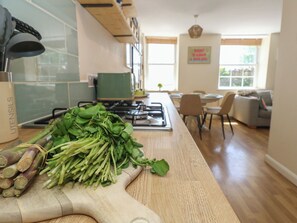 Una cocina con una tabla de cortar llena de vegetales de hoja y raíces, una estufa y un comedor con sillas y una mesa.