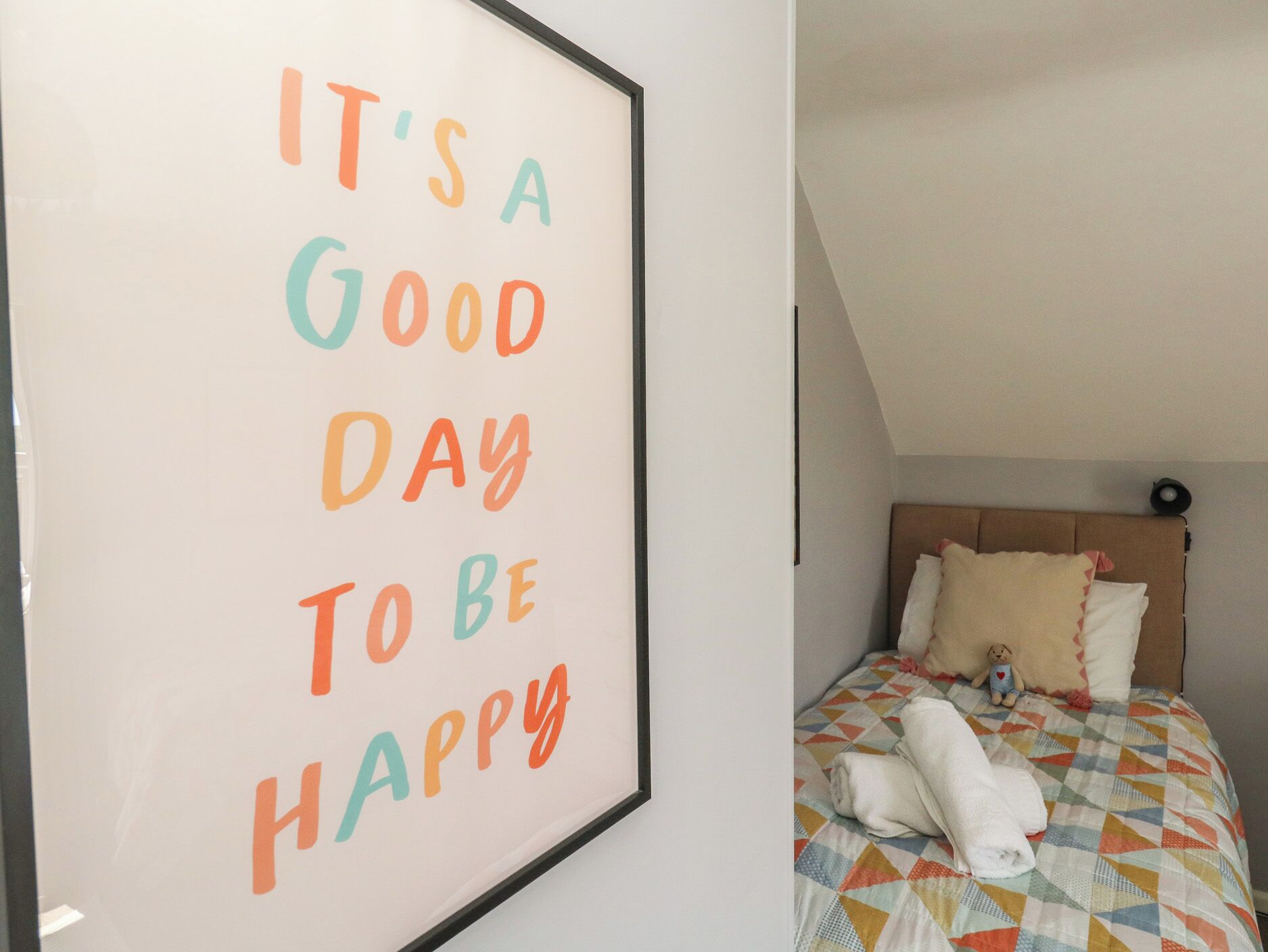 Una cita inspiradora enmarcada en una pared, una cama bien tendida con una colcha estampada y un cabecero con almohadones.