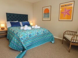 Habitación de hotel con una cama, una silla, una mesita de noche con lámpara y dos cuadros en la pared.