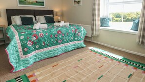 A bedroom with a bed, a window with curtains, a nightstand, and a wall-mounted lamp. - Mallow Cottage (Swanage)